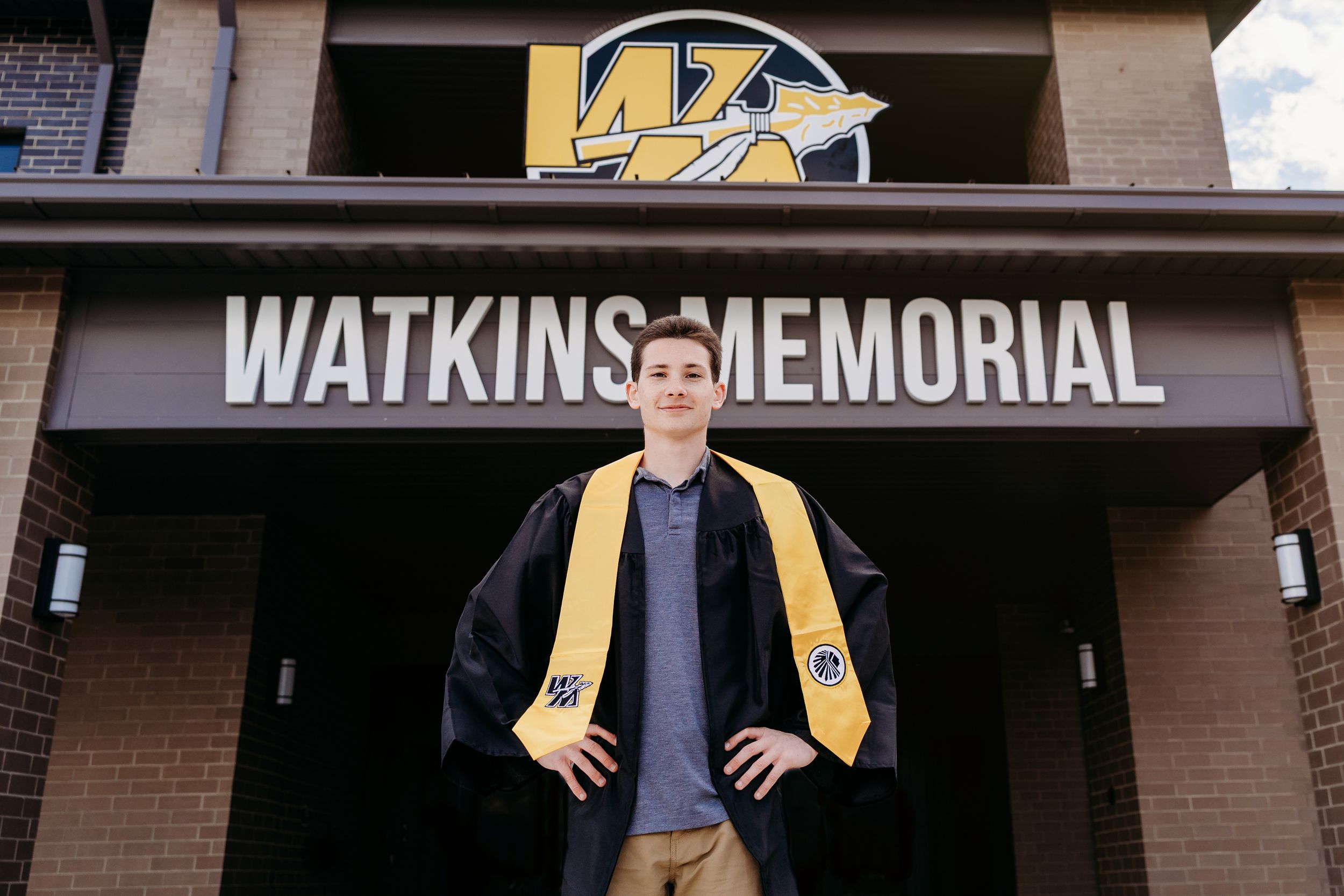 Graduate in cap and gown poses proudly in front of Watkins Memorial building wearing gold honor stole.