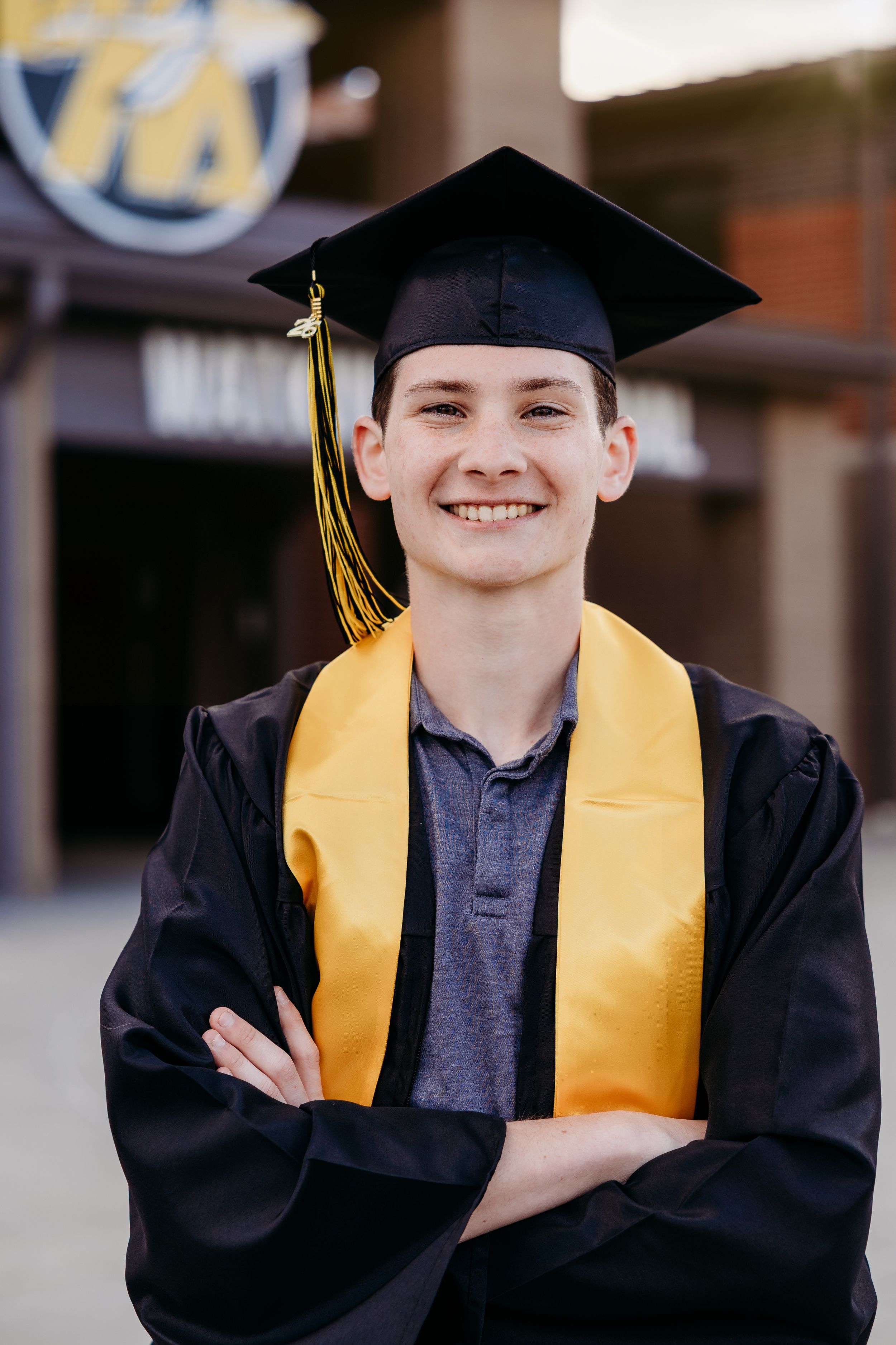 Smiling young graduate in black cap and gown with gold tassel and stole, arms crossed, posing outdoors.