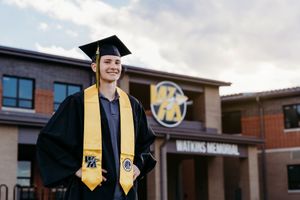 Graduate in cap and gown with gold stole smiles proudly in front of Watkins Memorial High School.