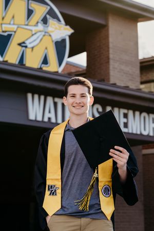 Smiling graduate in black cap and gold stole holds diploma outside Watkins Memorial building.