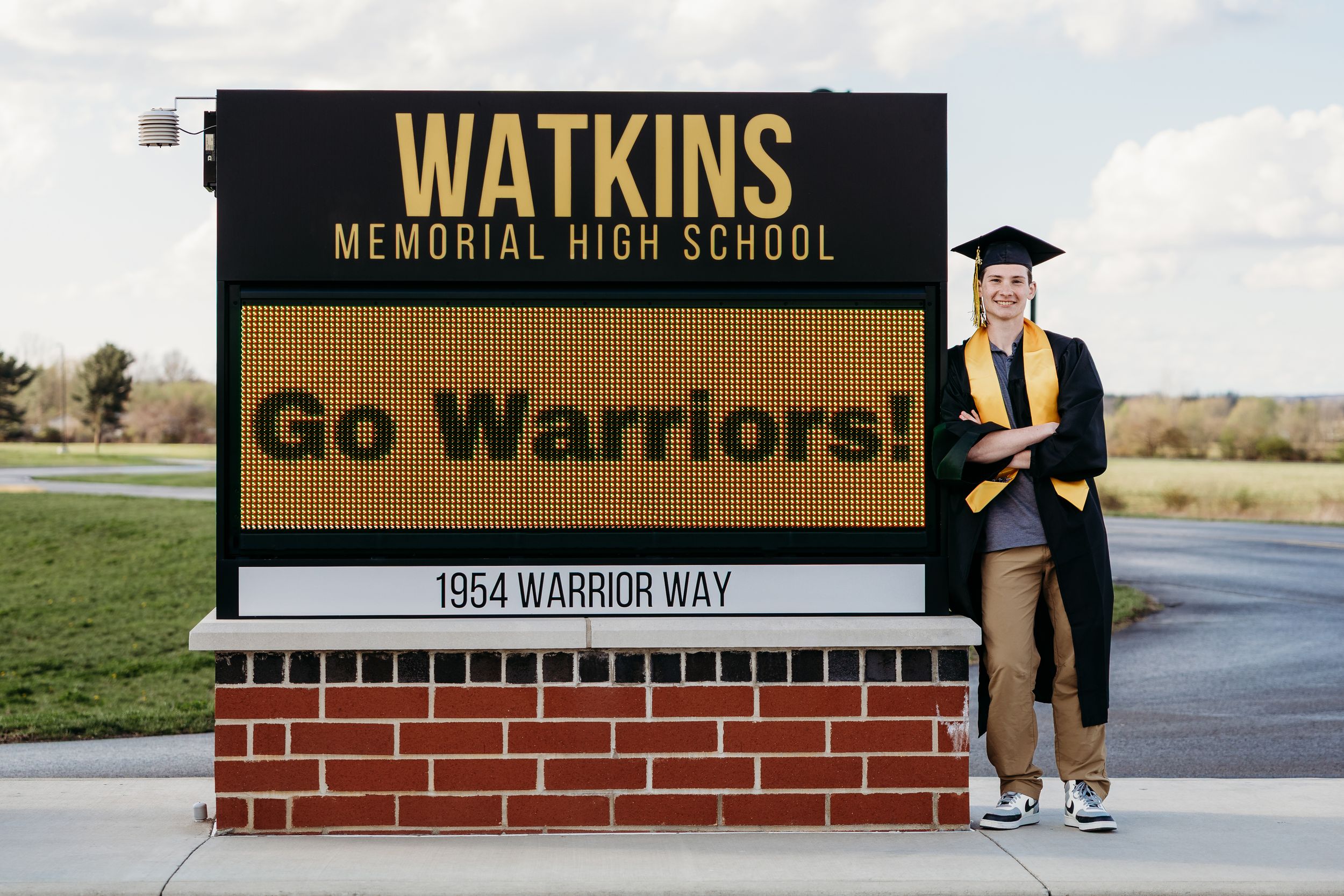 Graduate in cap and gown poses proudly beside Watkins Memorial High School sign reading Go Warriors.