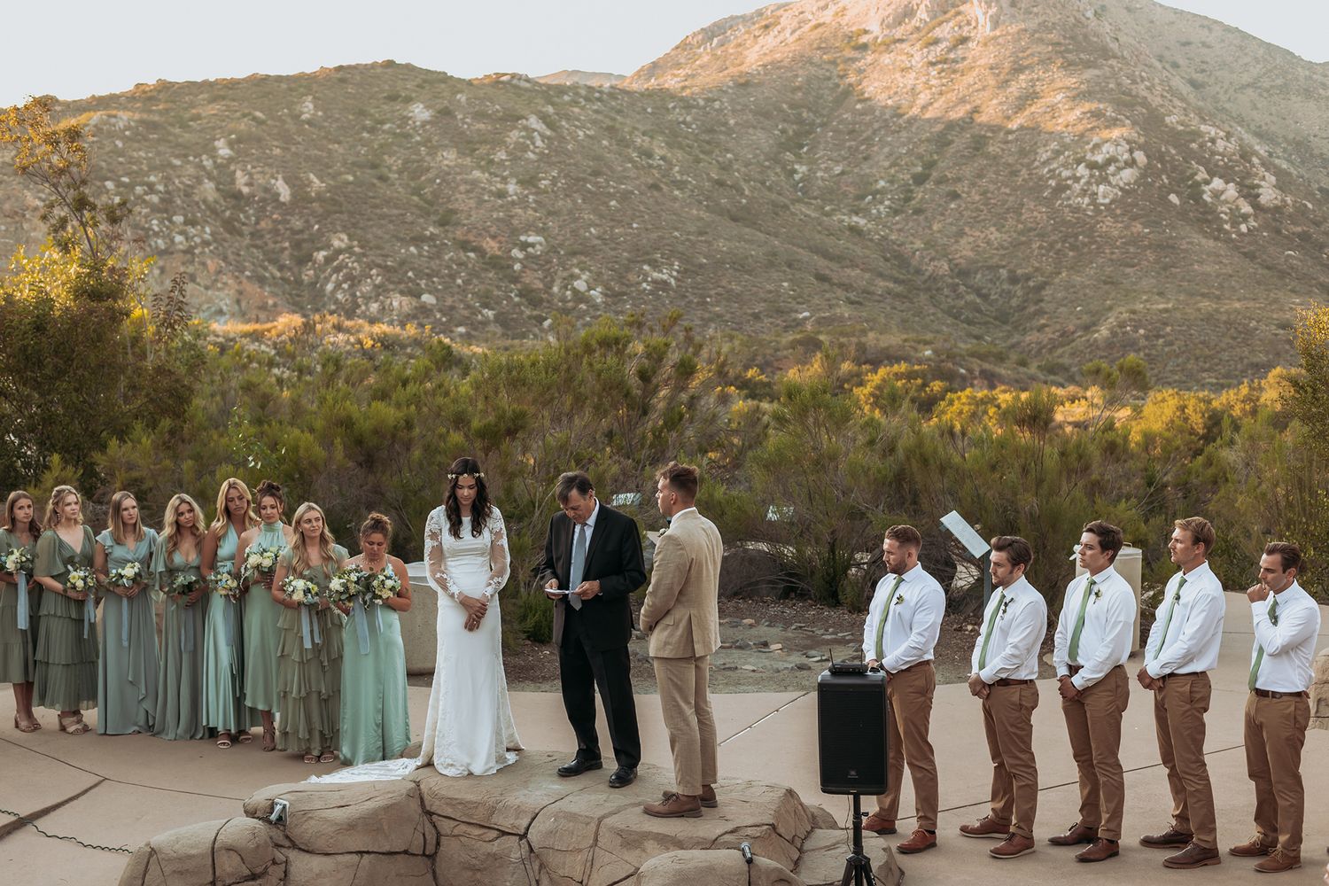 Outdoor wedding ceremony with bridesmaids in sage green dresses and groomsmen in tan suits, set against a scenic mountain backdrop.