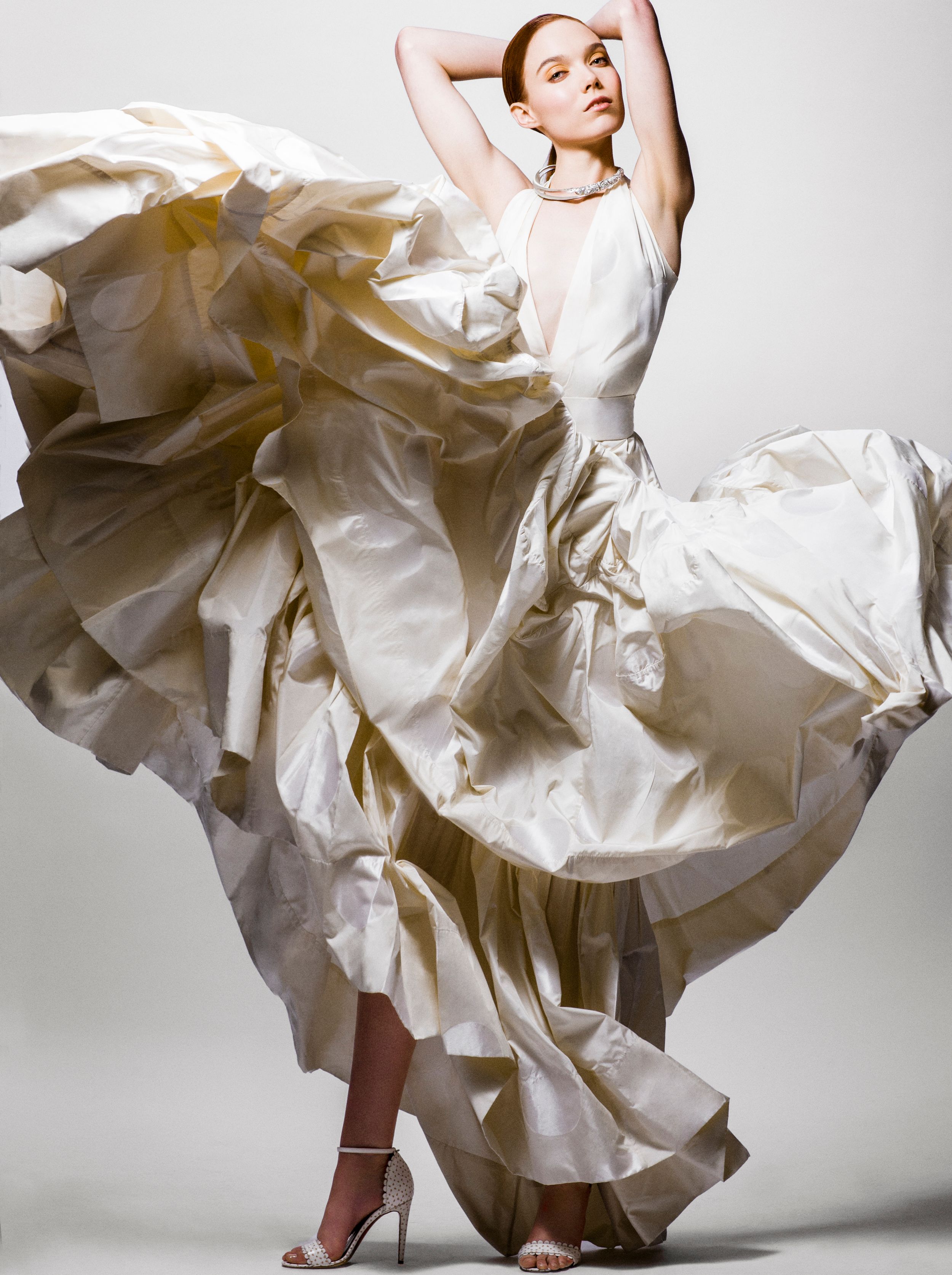 A model poses dynamically in a flowing ivory silk ballgown with deep neckline and silver heels against a white background.