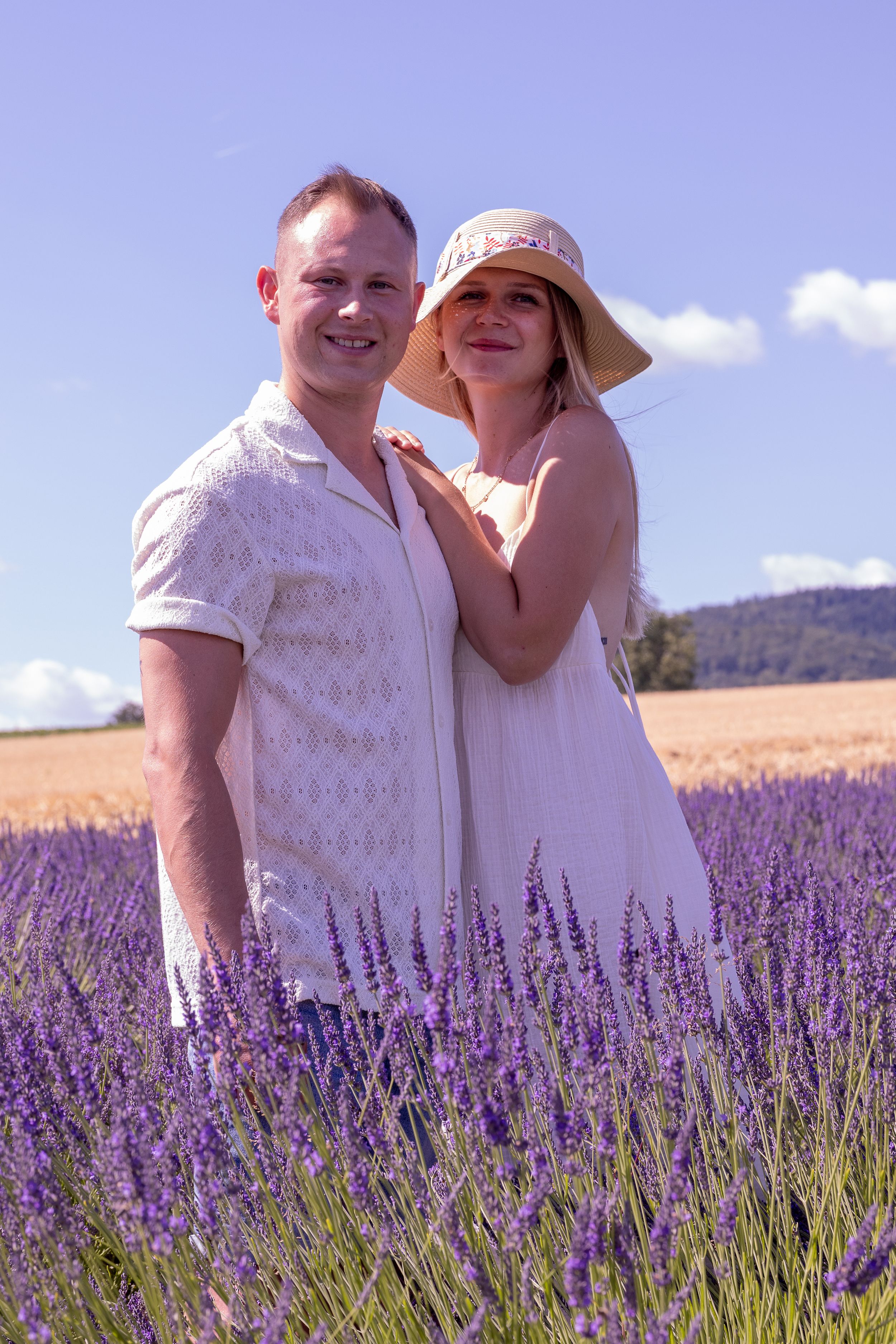 Couple embracing in a lavender field, woman in a hat smiling over man's shoulder on a sunny day.