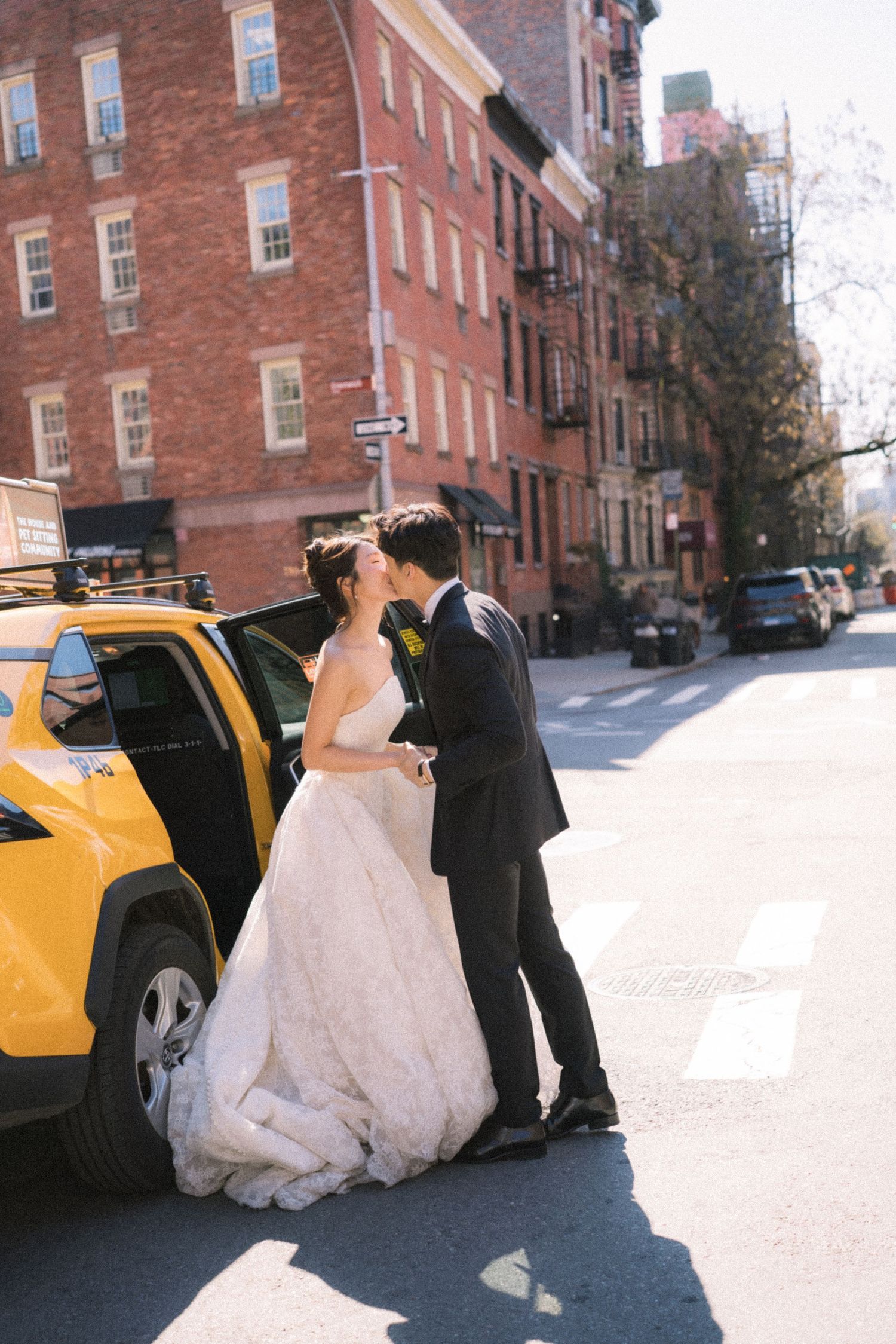 Bride and groom share a kiss beside a yellow NYC taxi cab on a sunny city street with brick buildings.