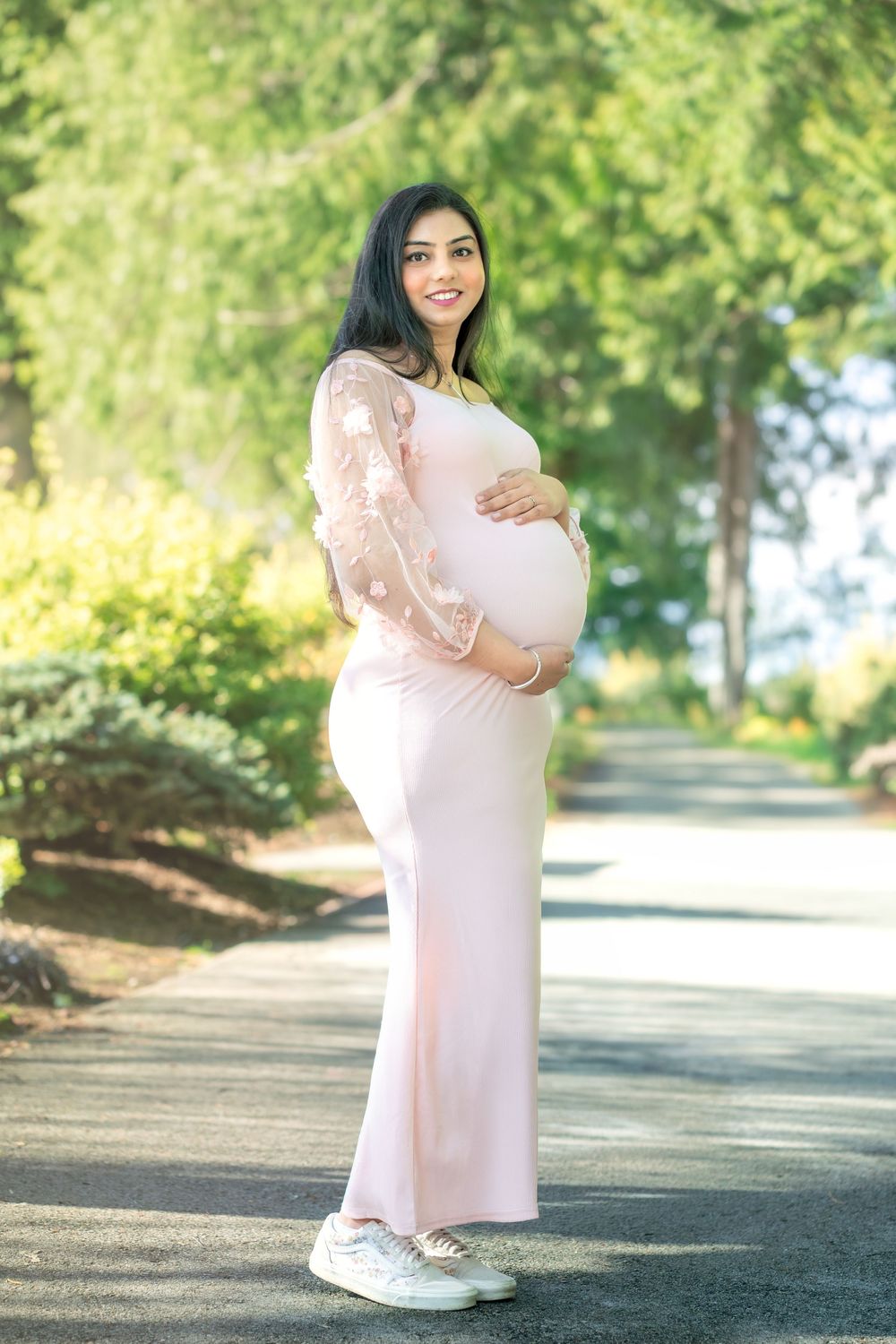 Smiling pregnant woman in pink lace dress cradling baby bump outdoors on a sunny tree-lined path.