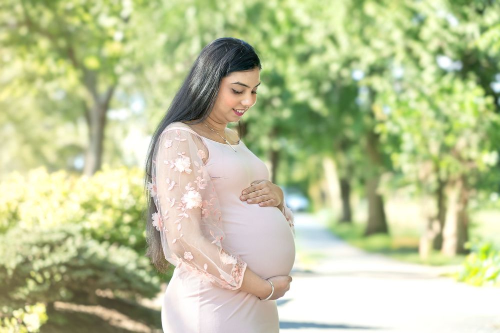 Pregnant woman in a blush pink lace gown cradling her baby bump outdoors in a sunlit green park.