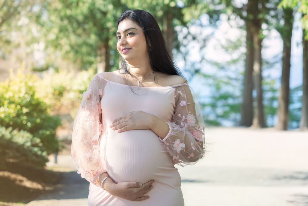Pregnant woman in pink dress cradling baby bump outdoors in sunny park setting.