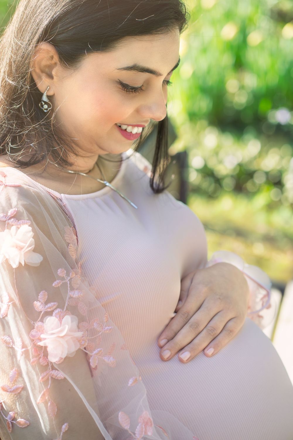 Smiling pregnant woman in floral pink dress cradling her baby bump outdoors in a sunlit garden.