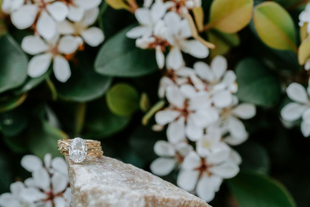 Gold engagement ring with oval diamond on stone, surrounded by white flowers and green leaves.