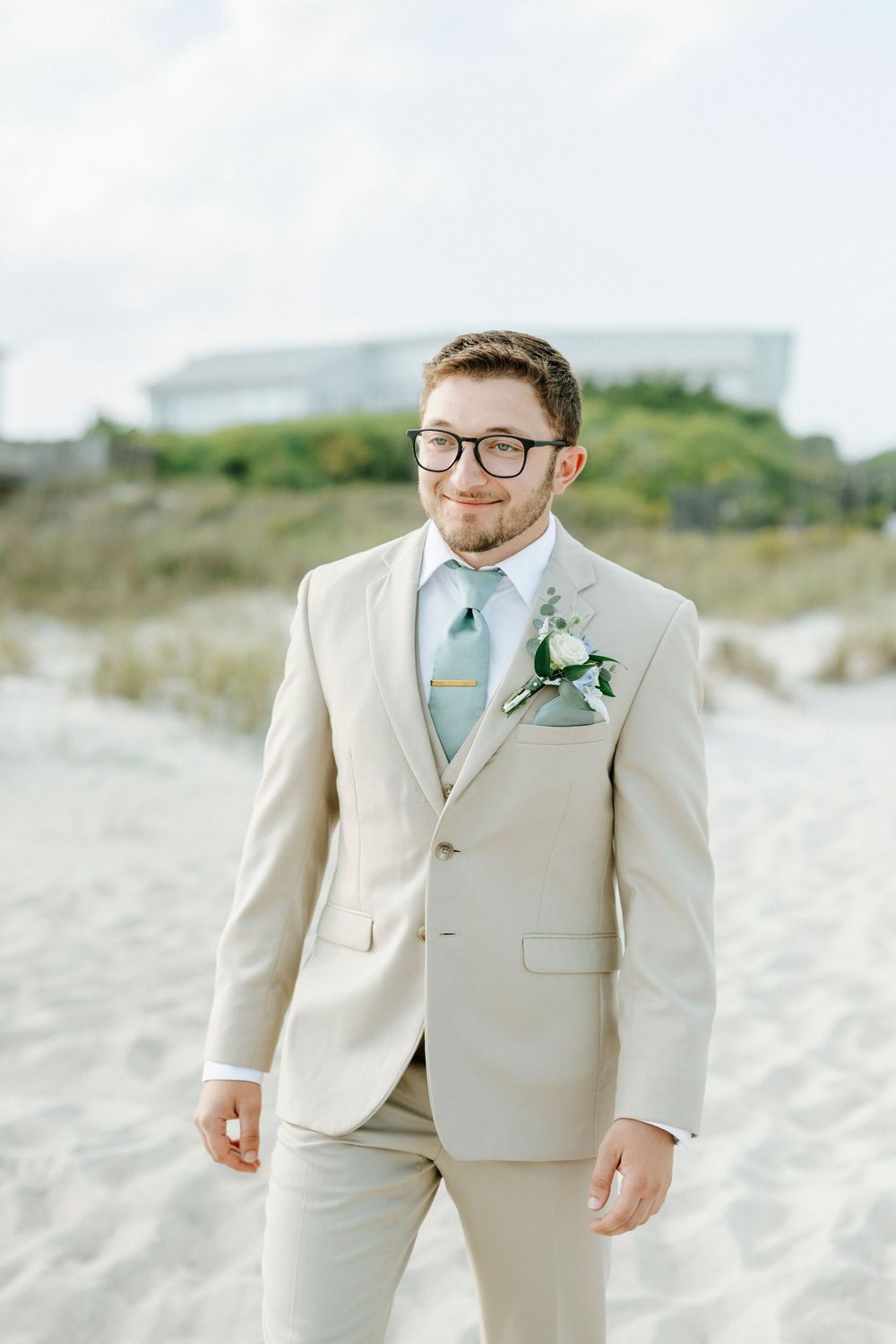 Groom in beige suit with mint tie and boutonniere standing on sandy beach during outdoor wedding.