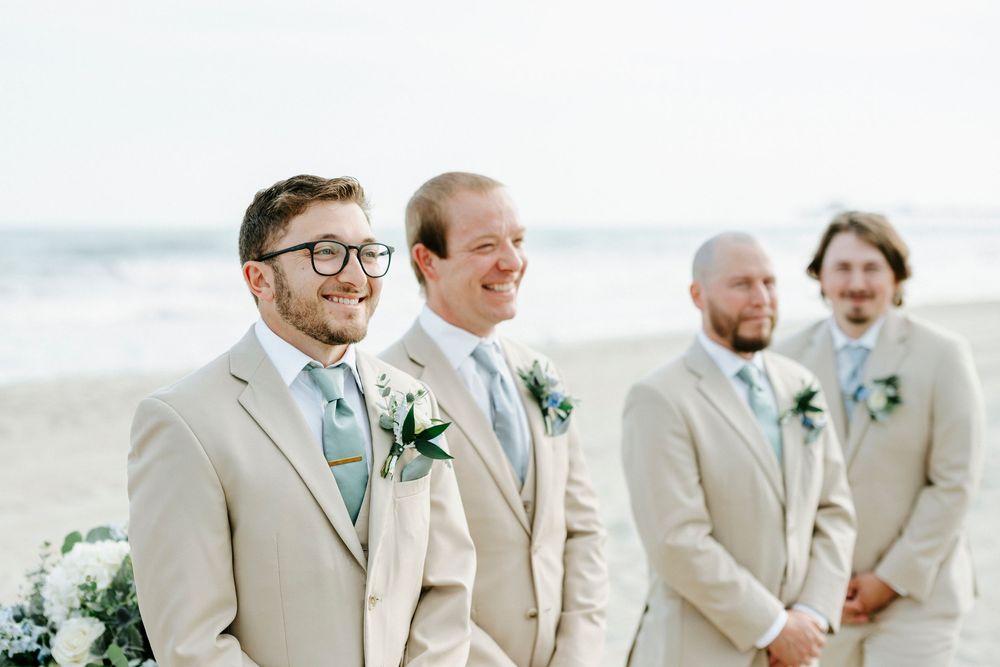 Four groomsmen in tan suits with sage green ties and boutonnieres smiling at a beach wedding ceremony.