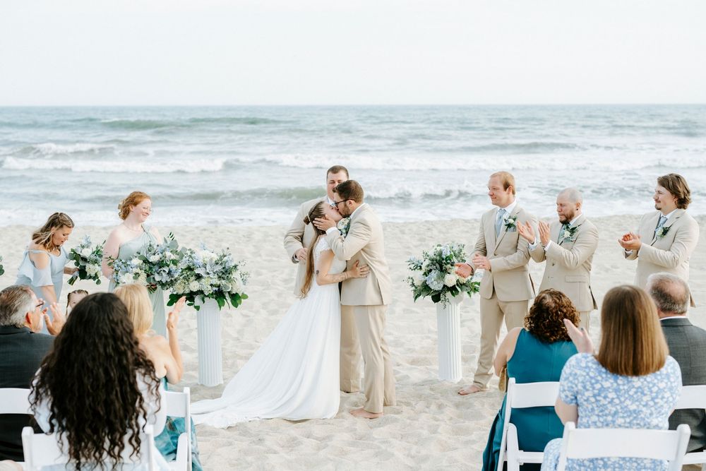 Bride and groom share first kiss at romantic beach wedding ceremony with ocean backdrop and white floral arrangements.