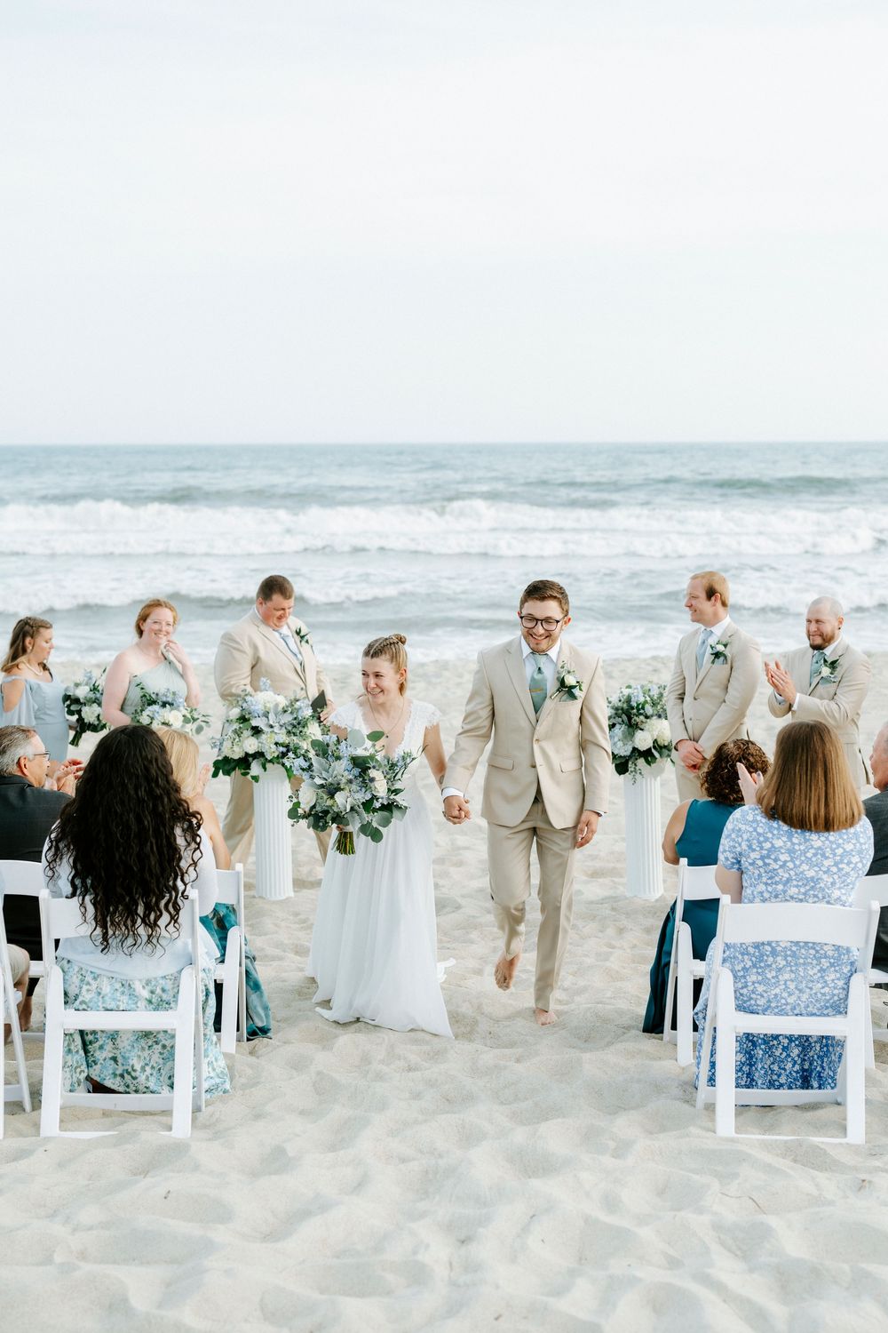 Bride and groom walk down sandy beach aisle hand-in-hand after oceanside wedding ceremony with guests seated on white chairs.