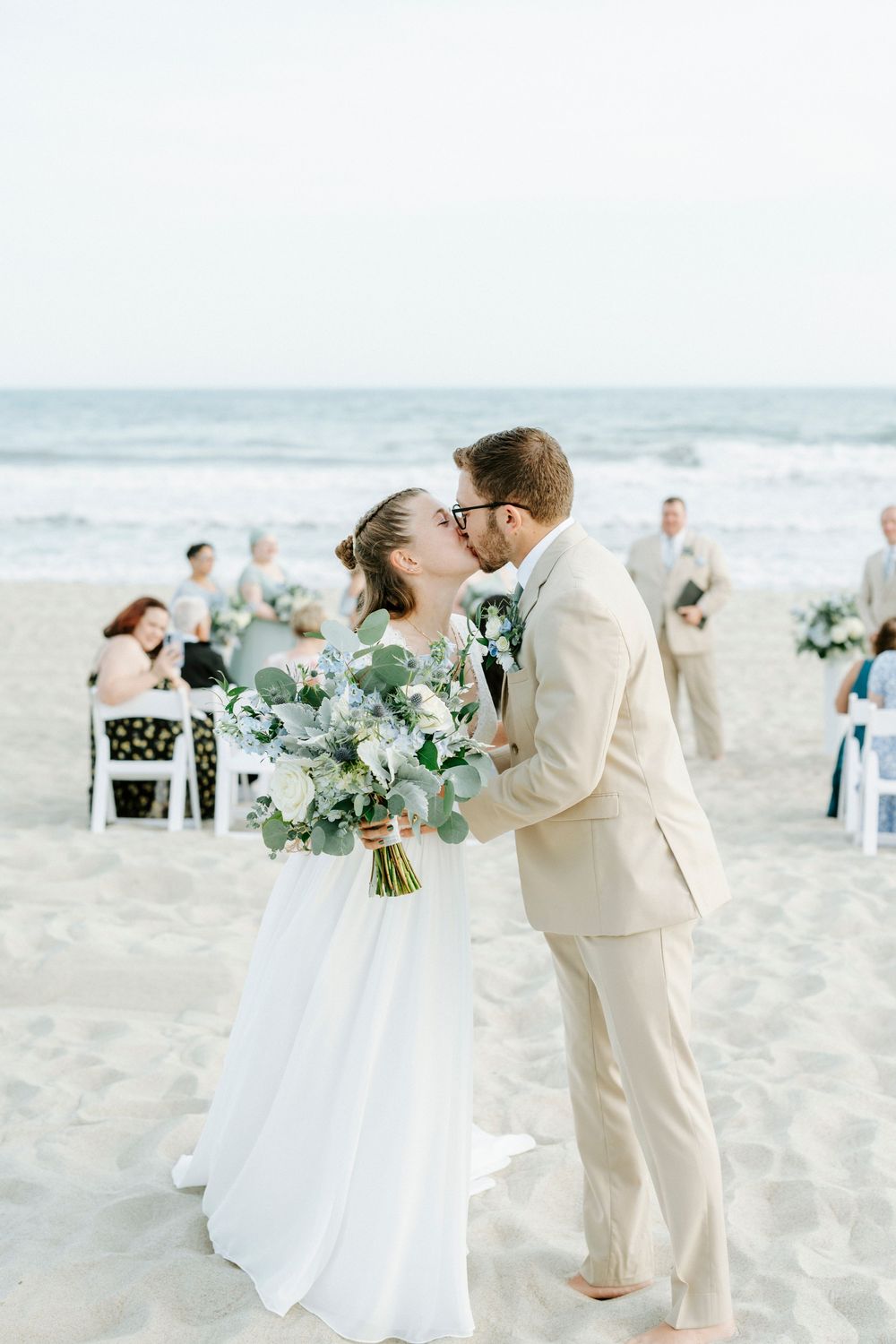 Bride and groom share first kiss on sandy beach with ocean backdrop and wedding guests seated behind.