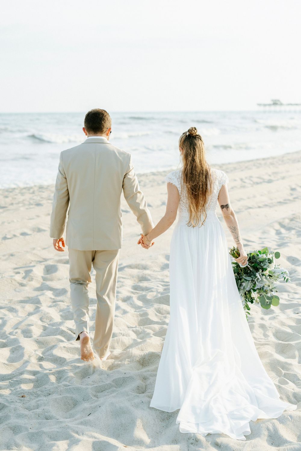 Bride in white gown and groom in beige suit walk hand in hand along sandy beach toward ocean.
