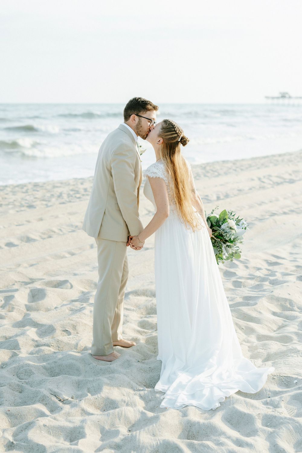 Bride and groom kissing on sandy beach, she holds greenery bouquet in white gown, he wears tan suit, ocean waves behind.