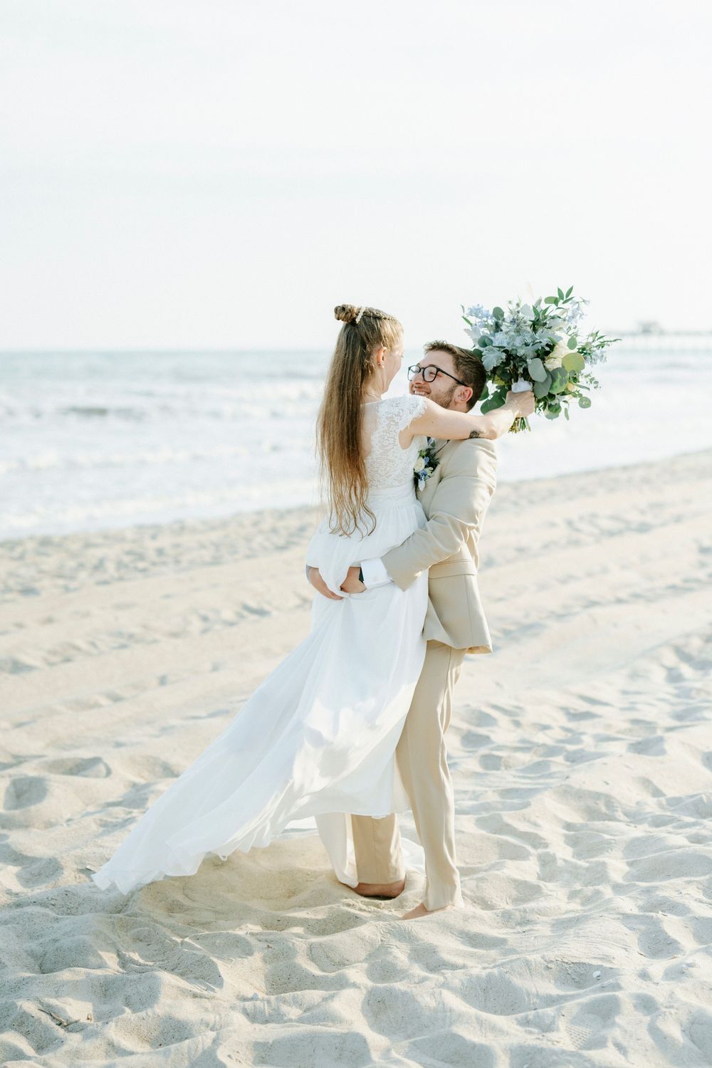 Groom lifting bride in white dress on sandy beach, holding floral bouquet with ocean waves in background.