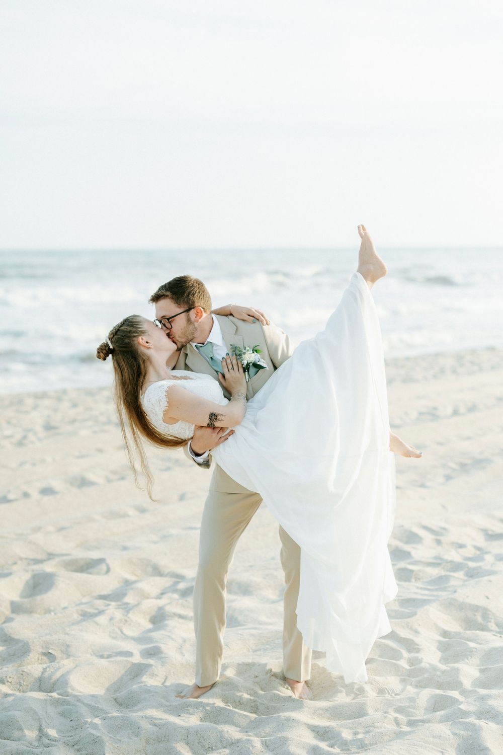 Groom lifts and dips bride for a romantic kiss on the sandy beach with ocean waves in the background.
