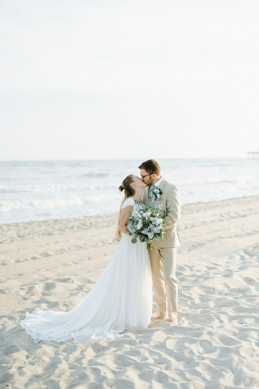 Bride and groom share a romantic moment on a sandy beach, with ocean waves and soft light in the background.