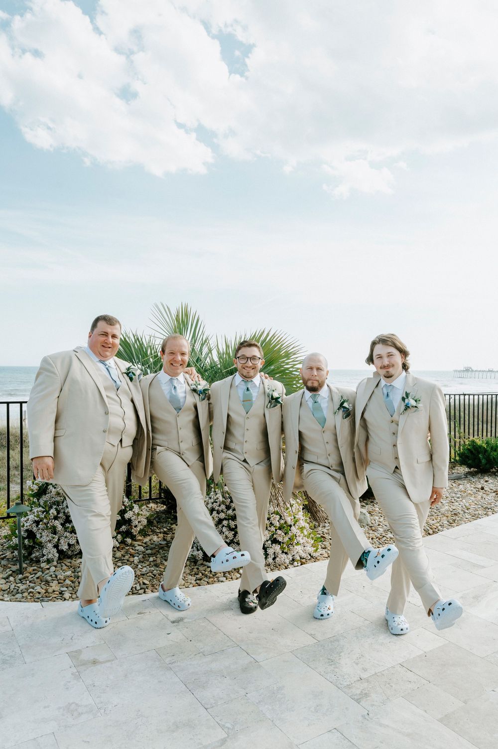 Five groomsmen in tan suits and blue Crocs posing outdoors with ocean backdrop.