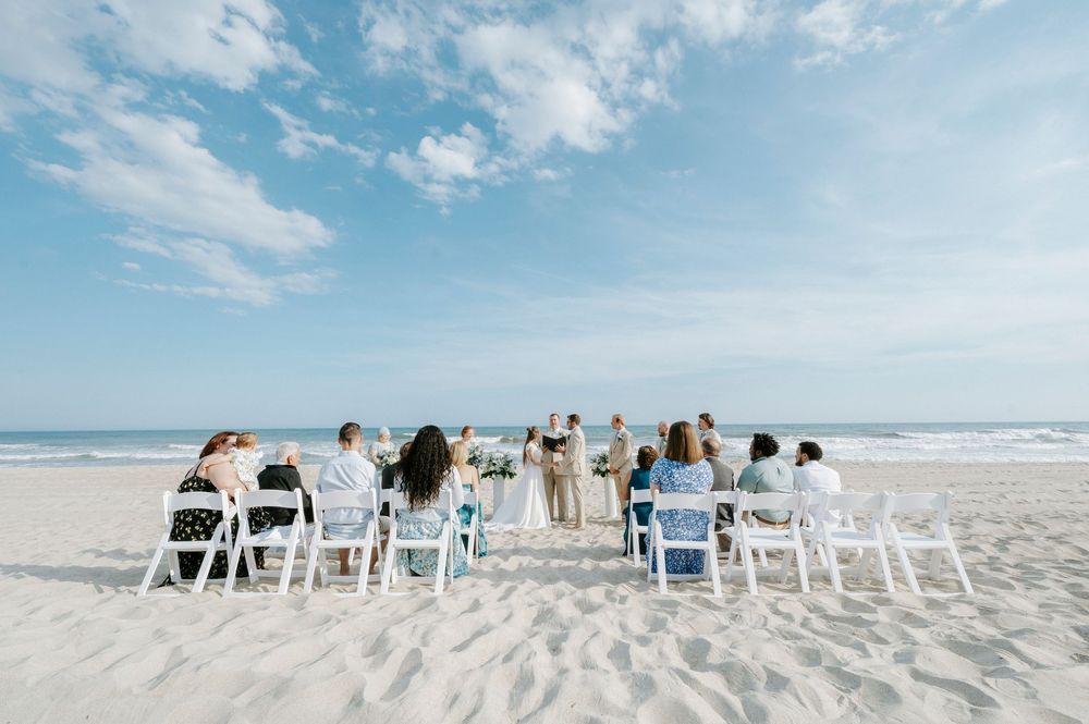 Small beach wedding ceremony with guests seated in white chairs facing the ocean under a blue sky.