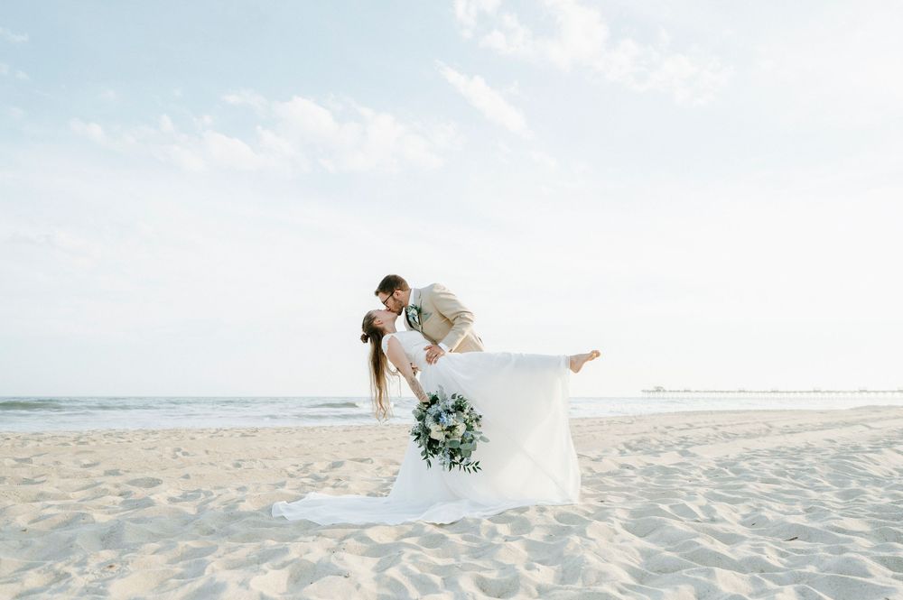 Couple sharing romantic dip kiss on sandy beach in wedding attire, bride holding blue bouquet.