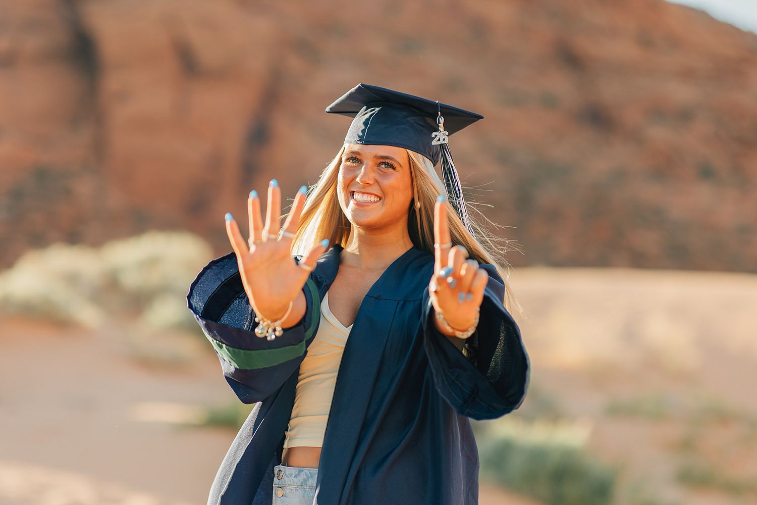 Smiling female graduate in cap and gown posing outdoors with hands raised showing seven fingers near red rock formations.