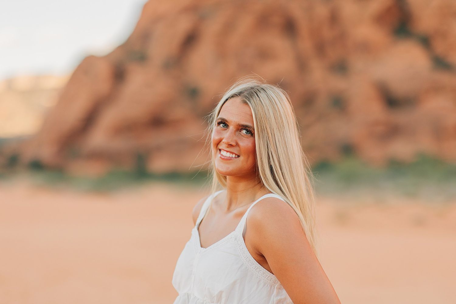 Young blonde woman in white sundress smiling outdoors with red rock formations in the desert background.
