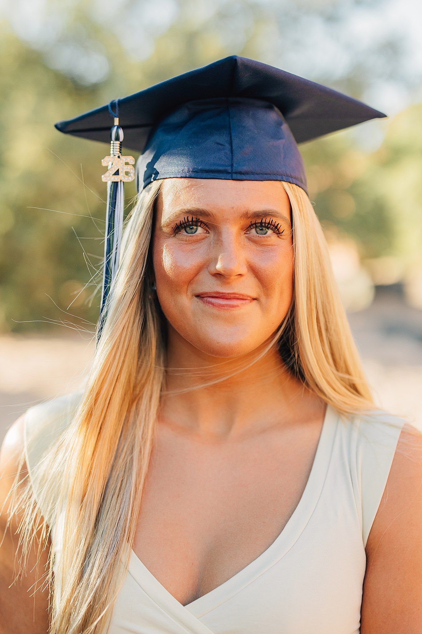 Smiling blonde woman in navy blue graduation cap with gold tassel and 26 charm, wearing white dress outdoors.
