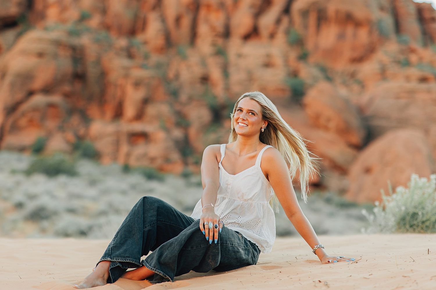 Smiling blonde woman sitting on desert sand with red rock formations in the background wearing jeans and white tank top.