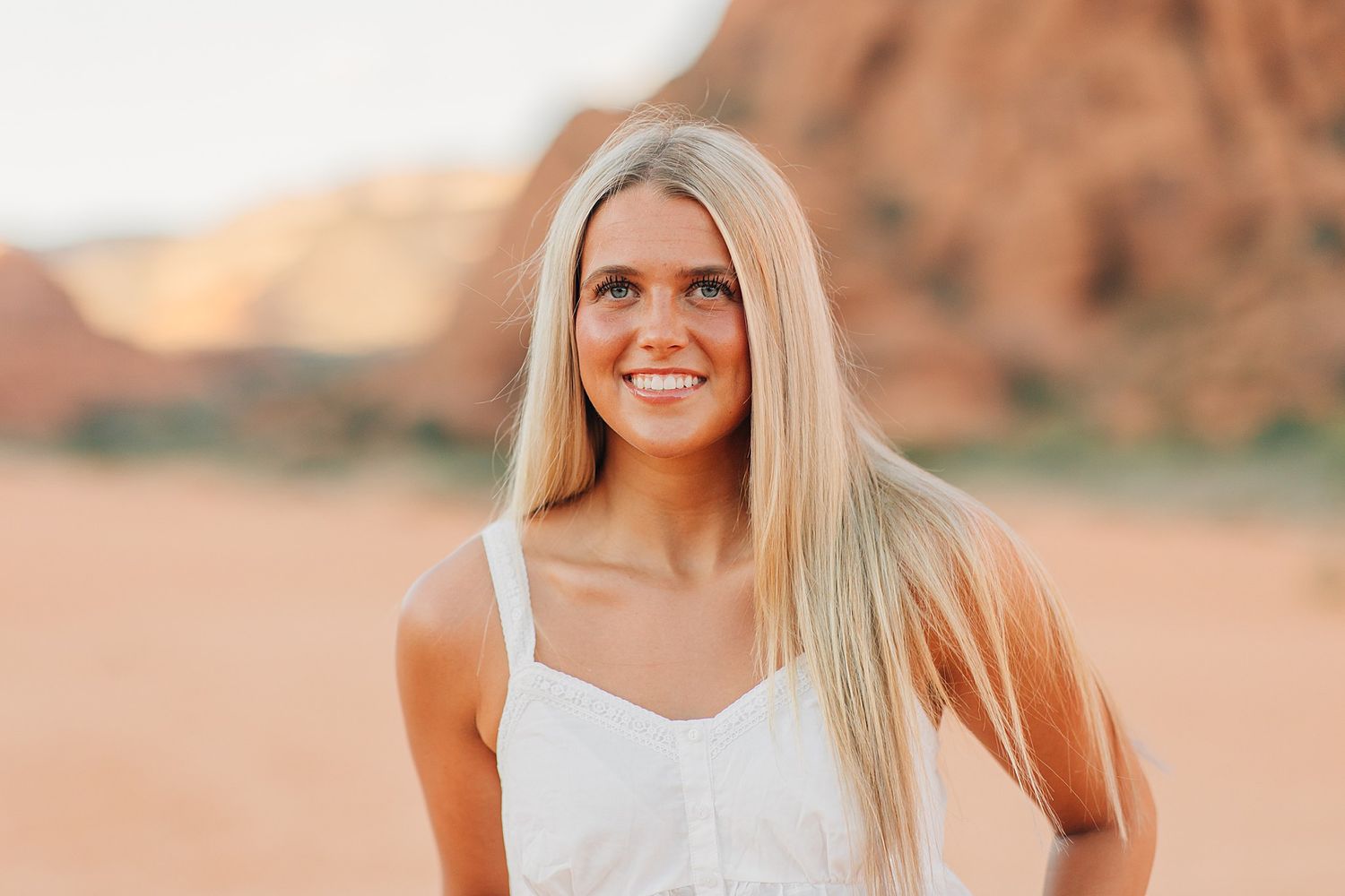 Smiling blonde woman in a white dress standing in a sunny red rock desert setting.