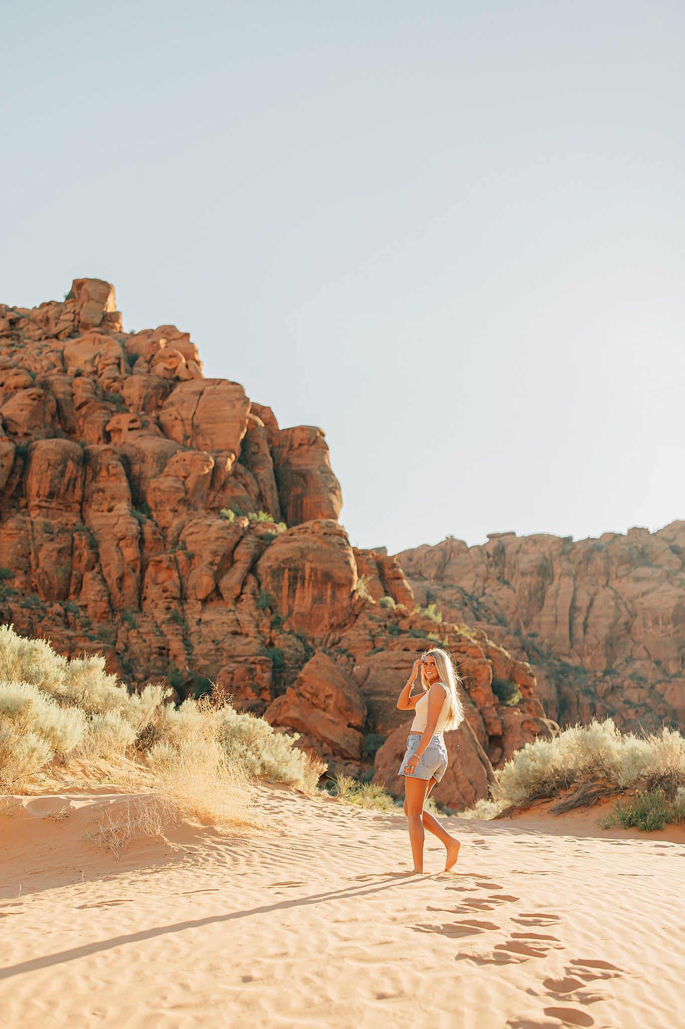 Woman walking on sandy desert dunes surrounded by dramatic red rock formations under a clear blue sky.