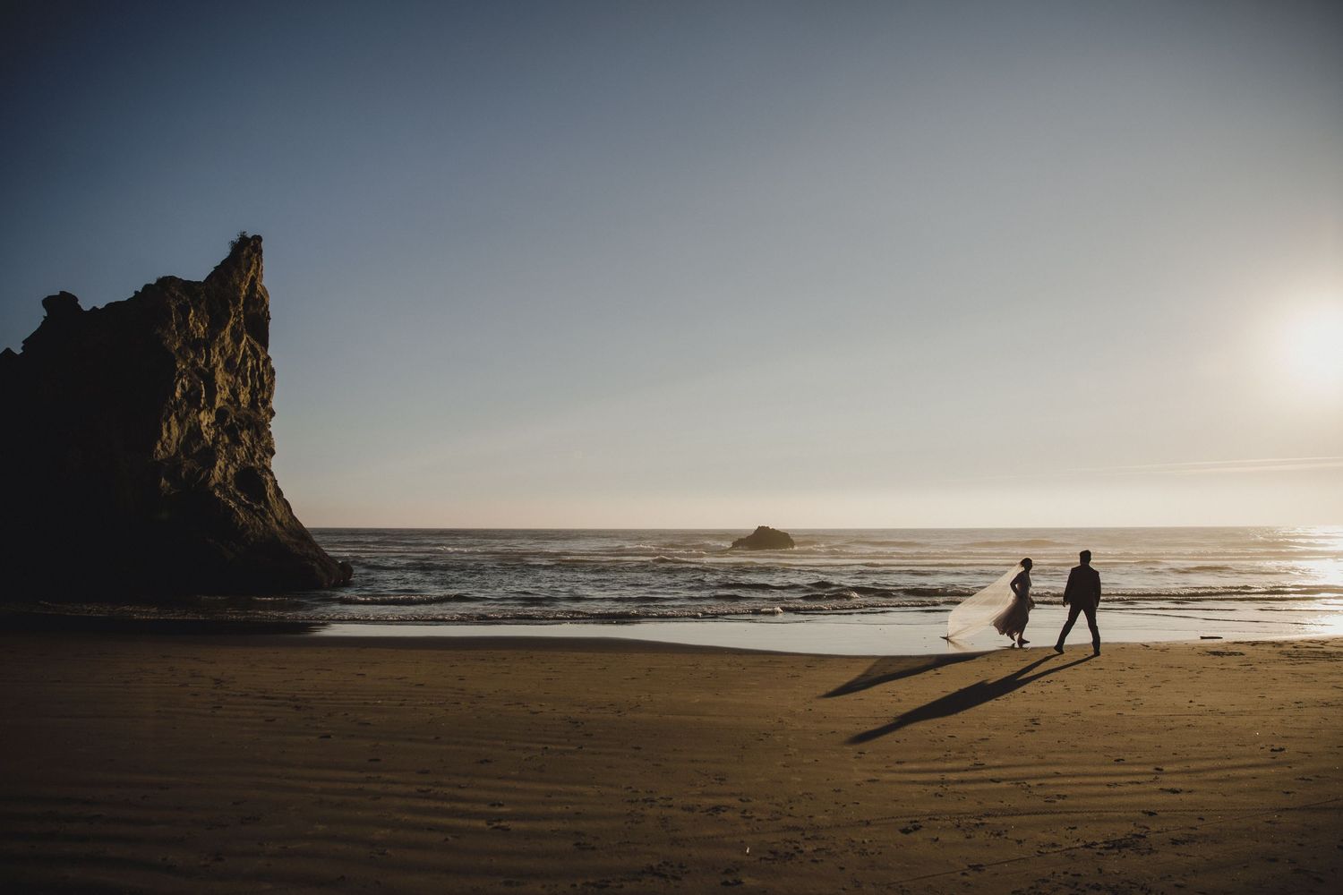 Bride and groom stand on coastal rocks with ocean waves and blue sky backdrop in a romantic beach wedding photo.
