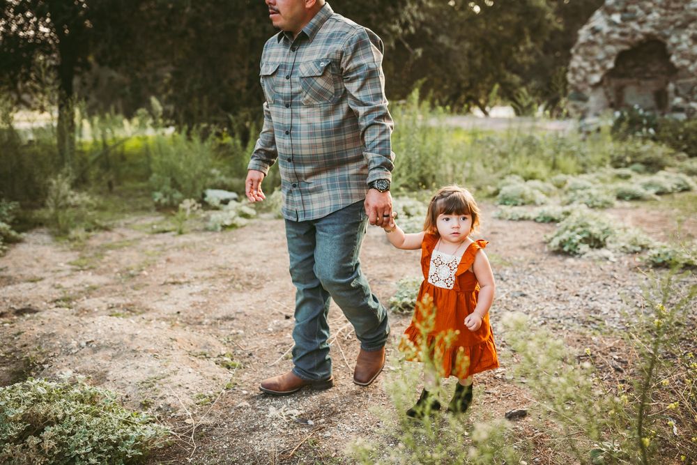 Father holds toddler girl's hand in rust orange dress while walking on a dirt path outdoors.