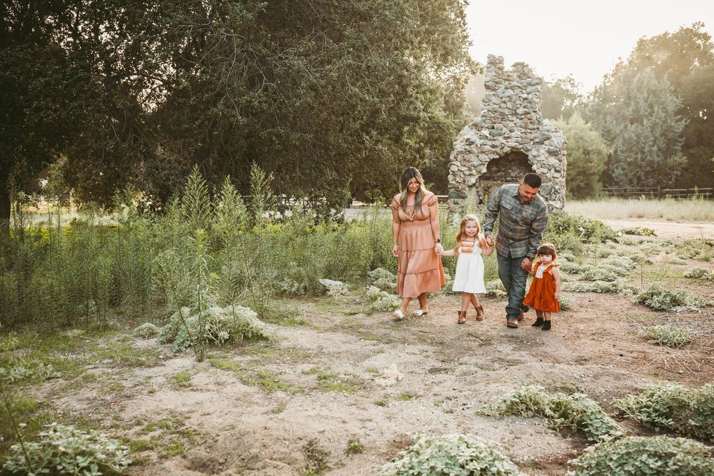 Family of four walking outdoors in a lush green park, parents with two young daughters in warm earthy tones.