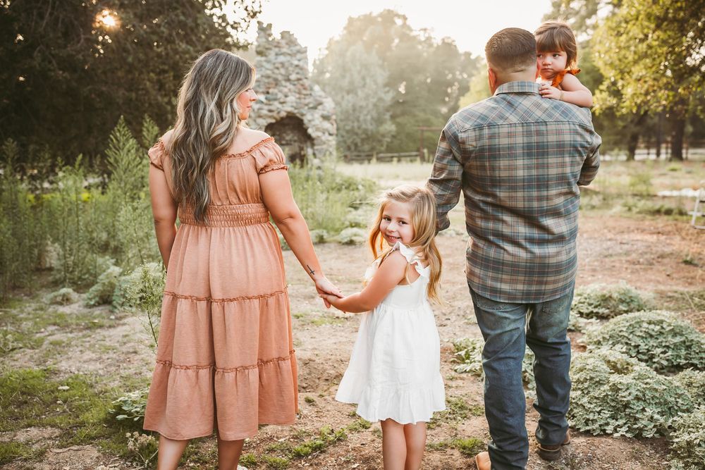 Family of four walking through a sunlit garden, parents holding hands with young daughter in white dress at golden hour.
