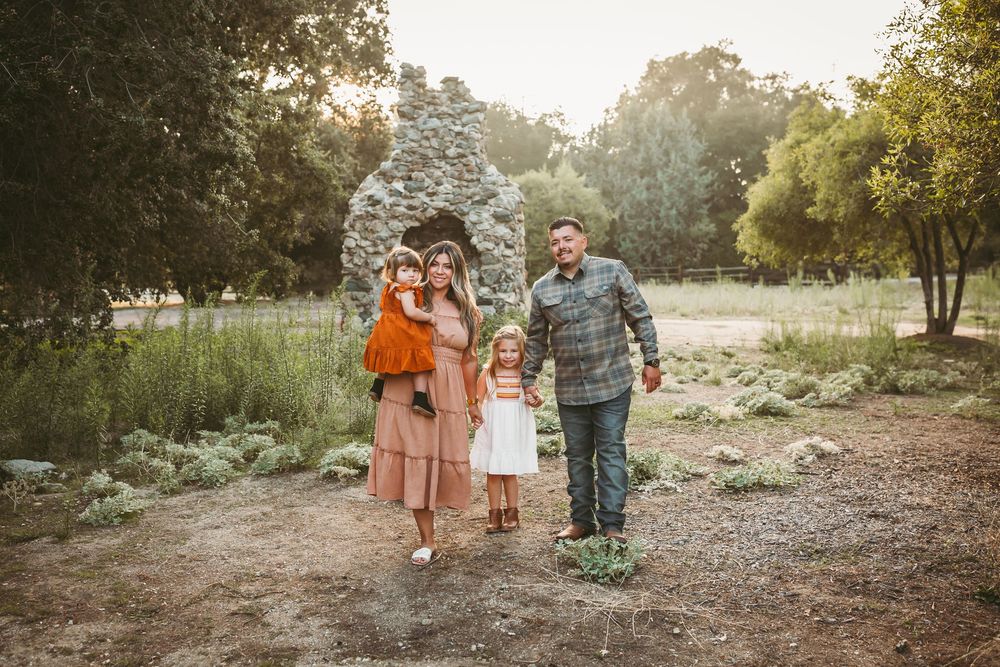 Family of four posing outdoors near a stone structure surrounded by lush greenery during golden hour.