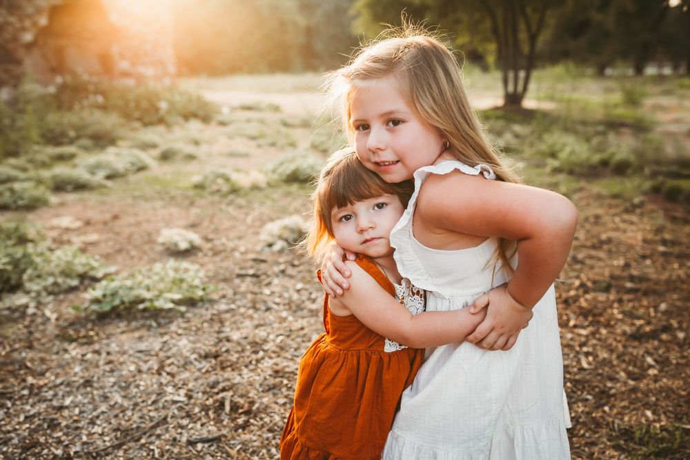 Two young girls hugging outdoors in warm golden sunlight, one in white dress, one in orange.