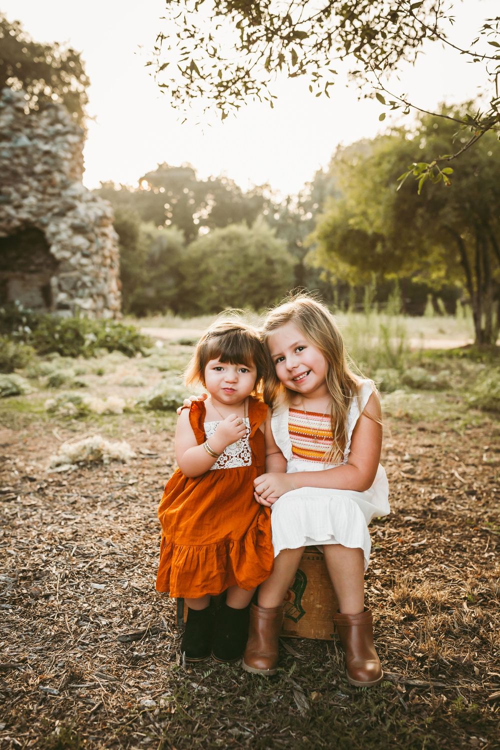Two young sisters in white and rust orange dresses sit together outdoors in a golden sunlit garden.