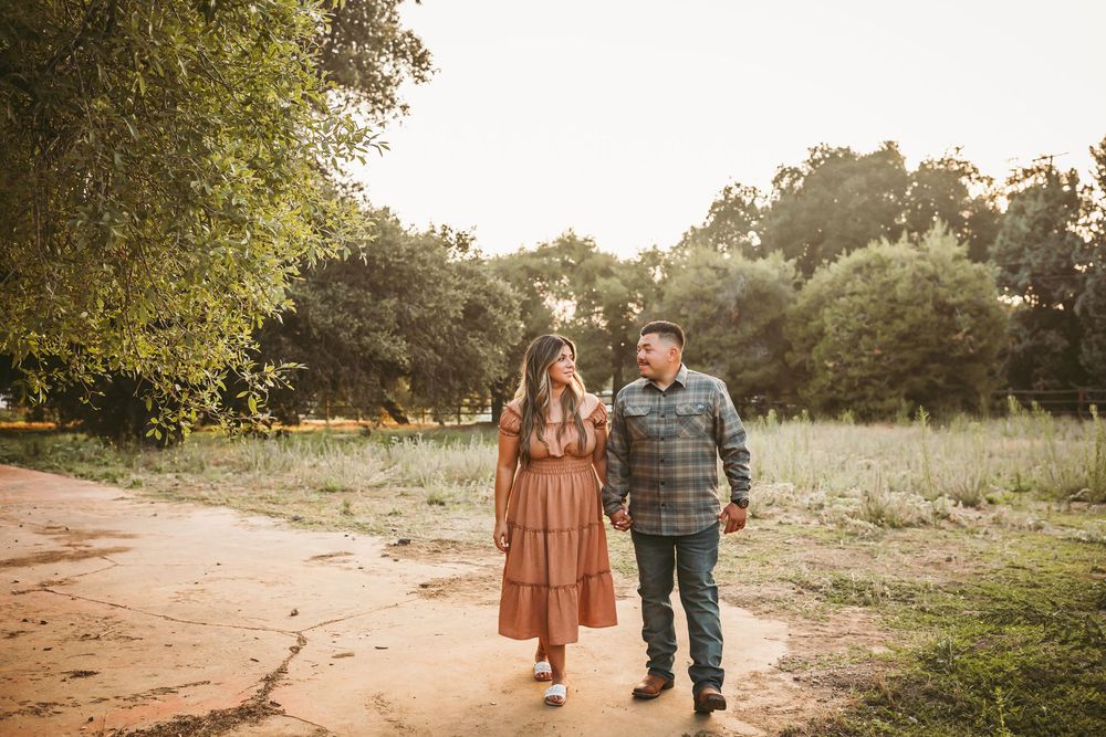 Couple walking hand in hand on a dirt path surrounded by lush green trees during golden hour.