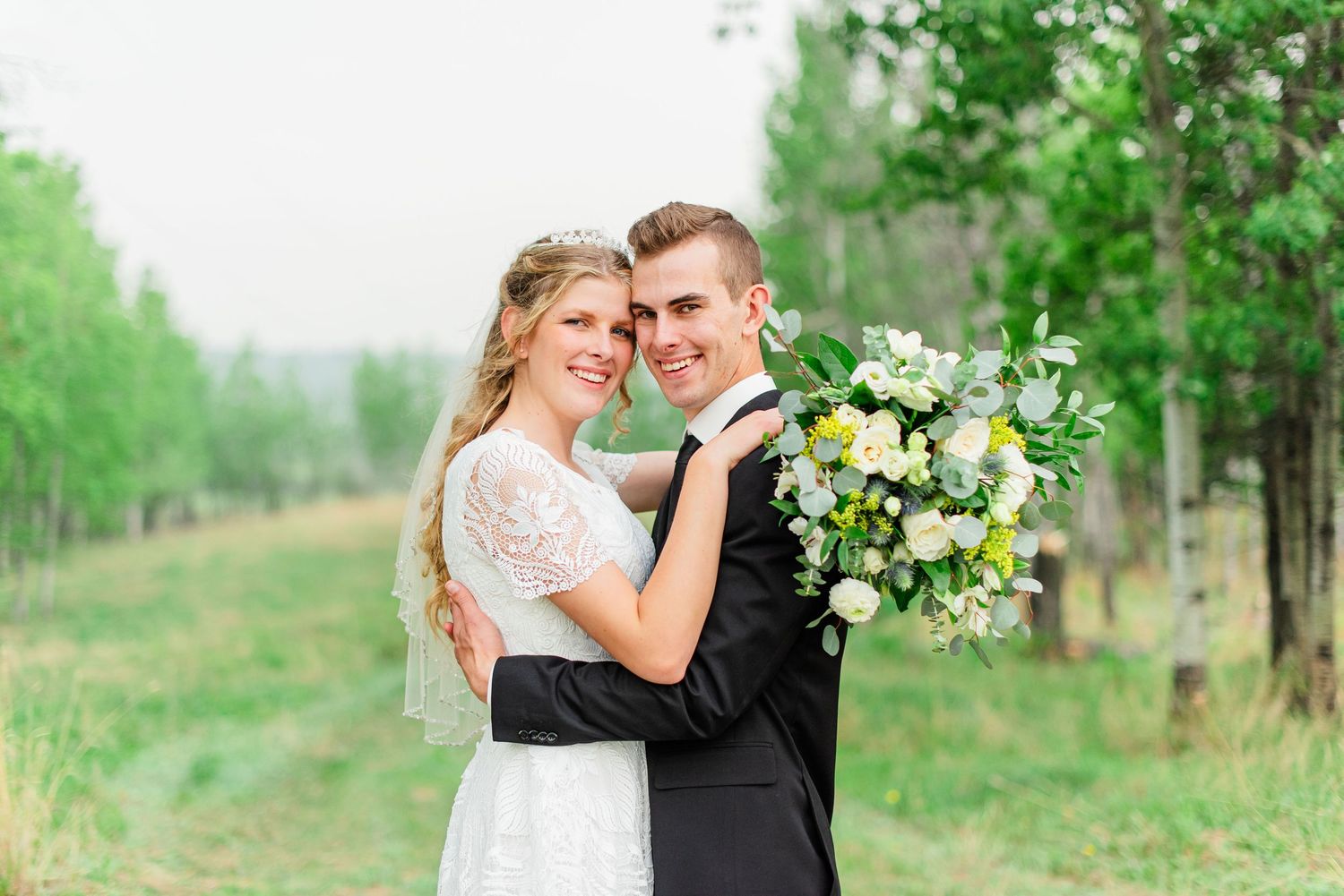 Bride and groom embracing among tall evergreen trees in a forest wedding portrait with light snow on the ground.