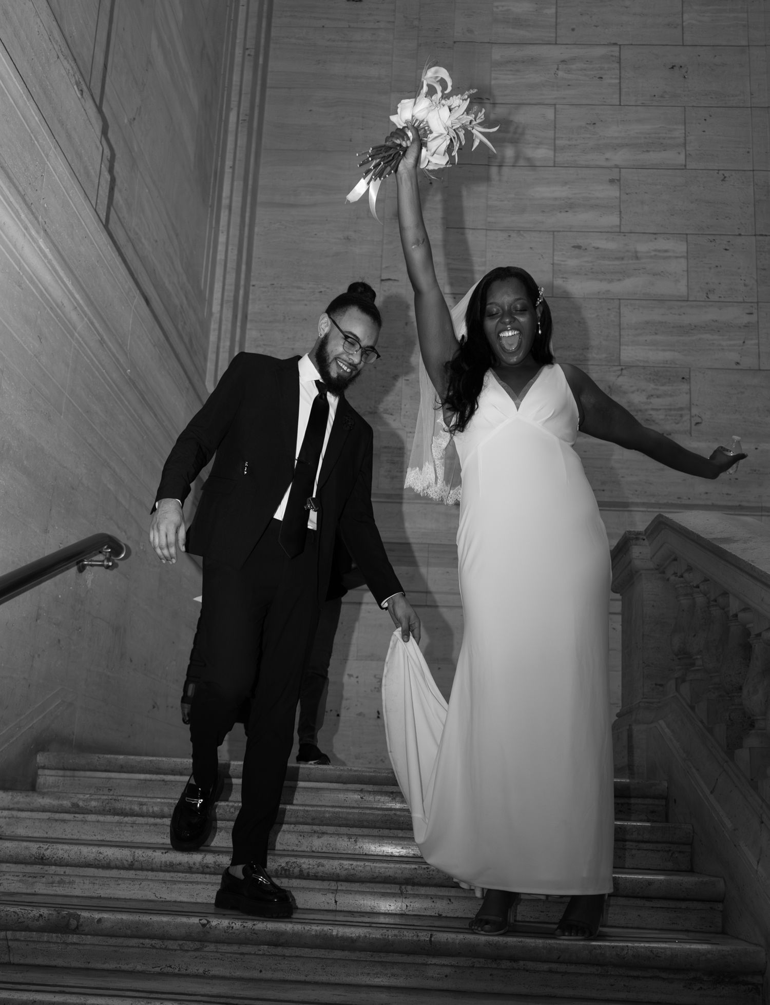 Black and white photo of a joyful newlywed couple celebrating on grand staircase, bride in white gown raising bouquet.