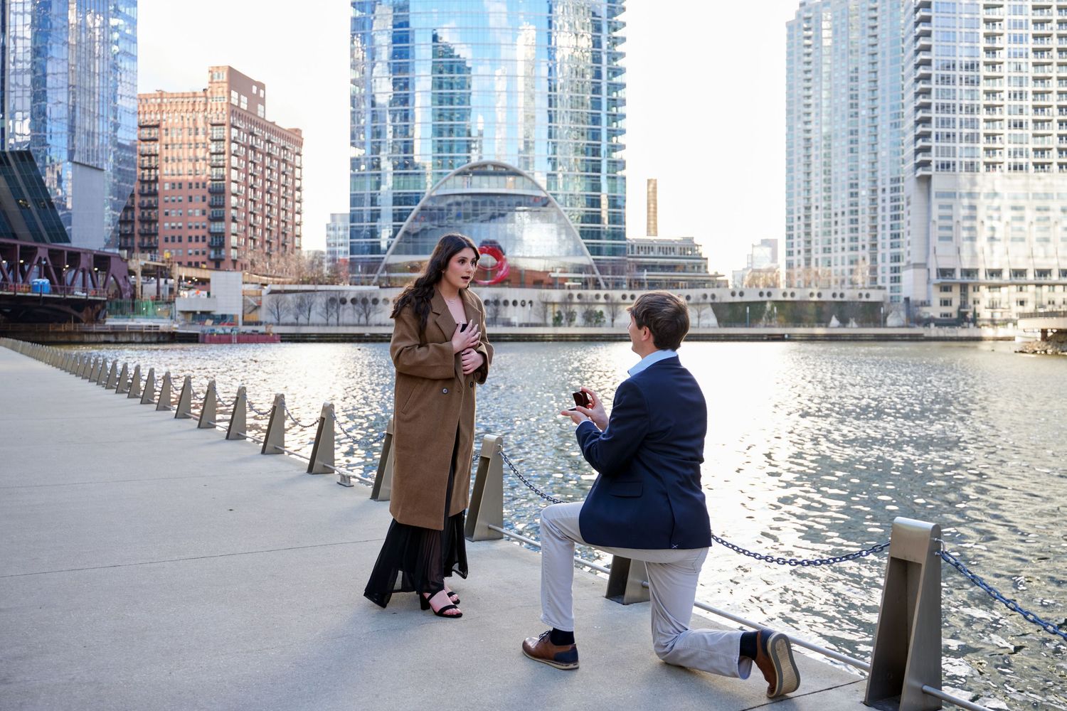 Man proposes to woman on one knee by Chicago River waterfront with city skyline in background.