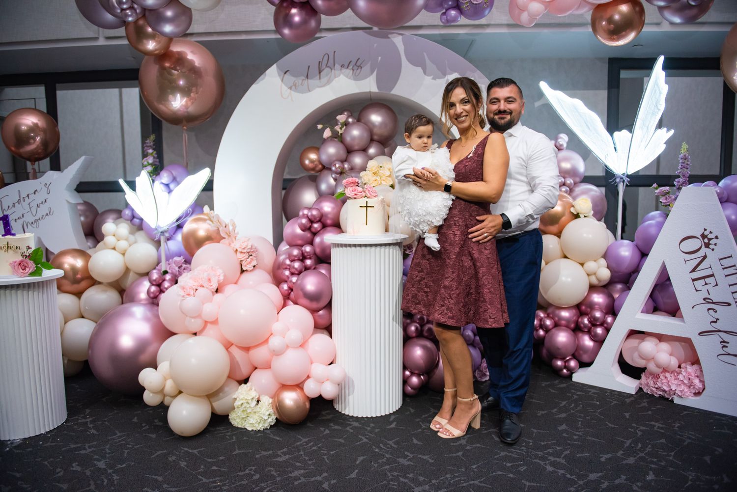 Family celebrating baby baptism with pink and rose gold balloon arch decor at reception venue.