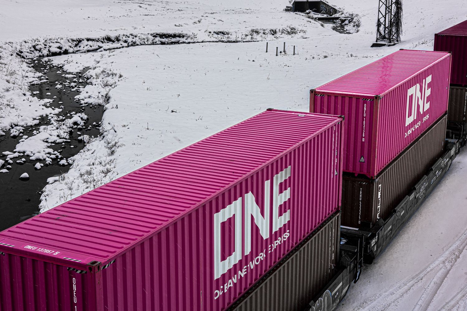 Two pink ONE shipping containers on a train traveling through a snowy winter landscape.