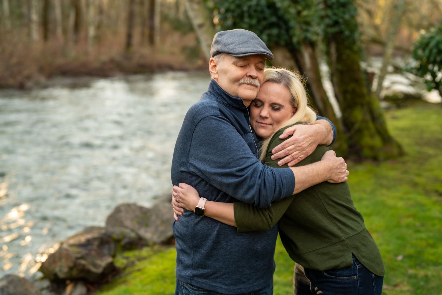 Candid family embraces by riverside with mossy rocks and trees.