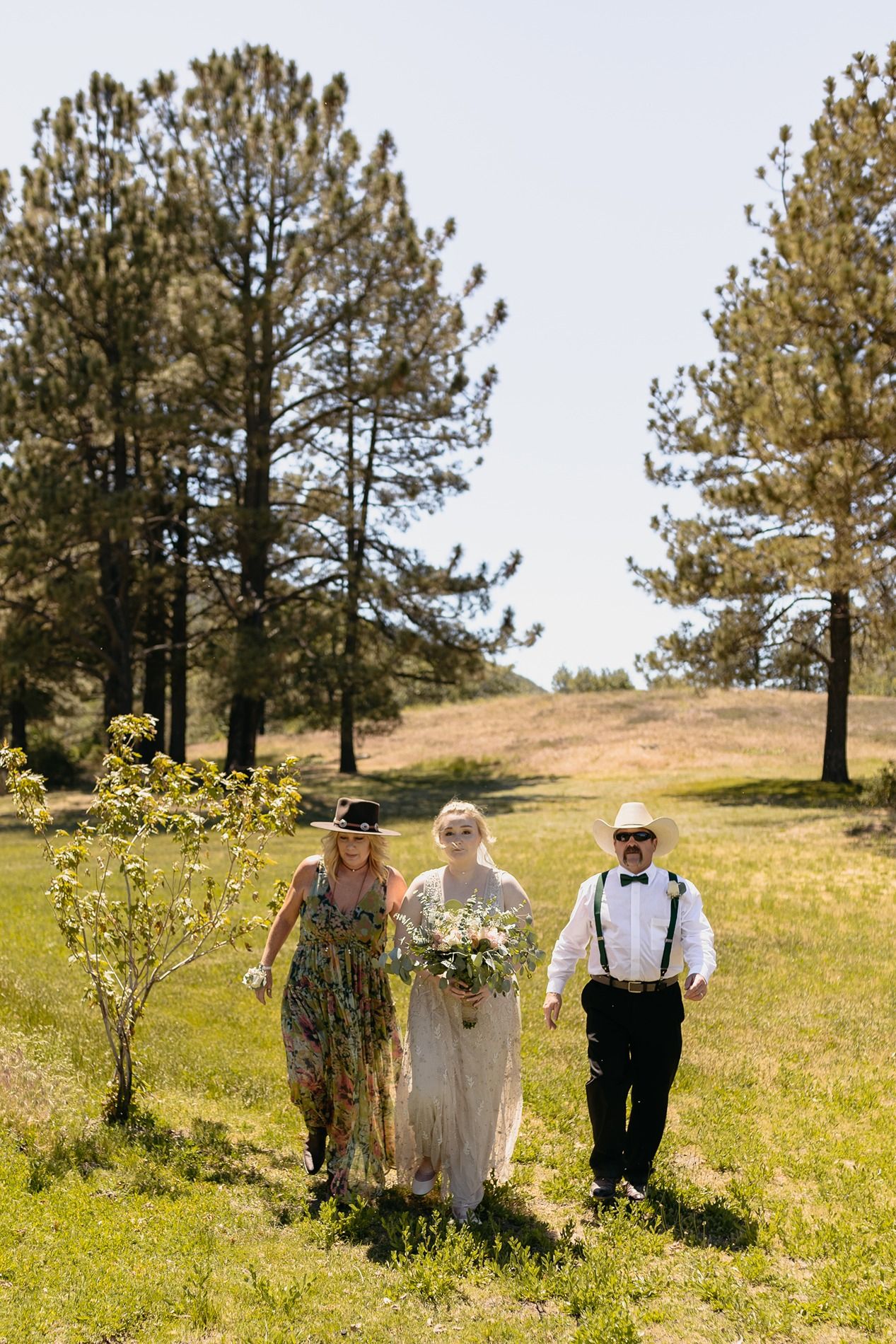 Bride walks down grassy outdoor aisle with parents amid tall pine trees on a sunny wedding day at Lake Cuyamaca, Julian.
