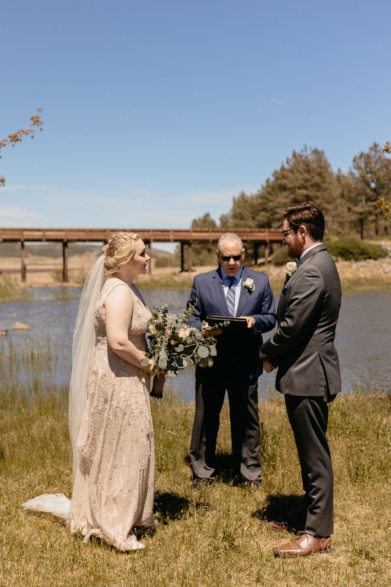 Outdoor wedding ceremony by a river with bride in sequin gown, groom in gray suit, and officiant holding a book at Lake Cuyamaca, Julian.