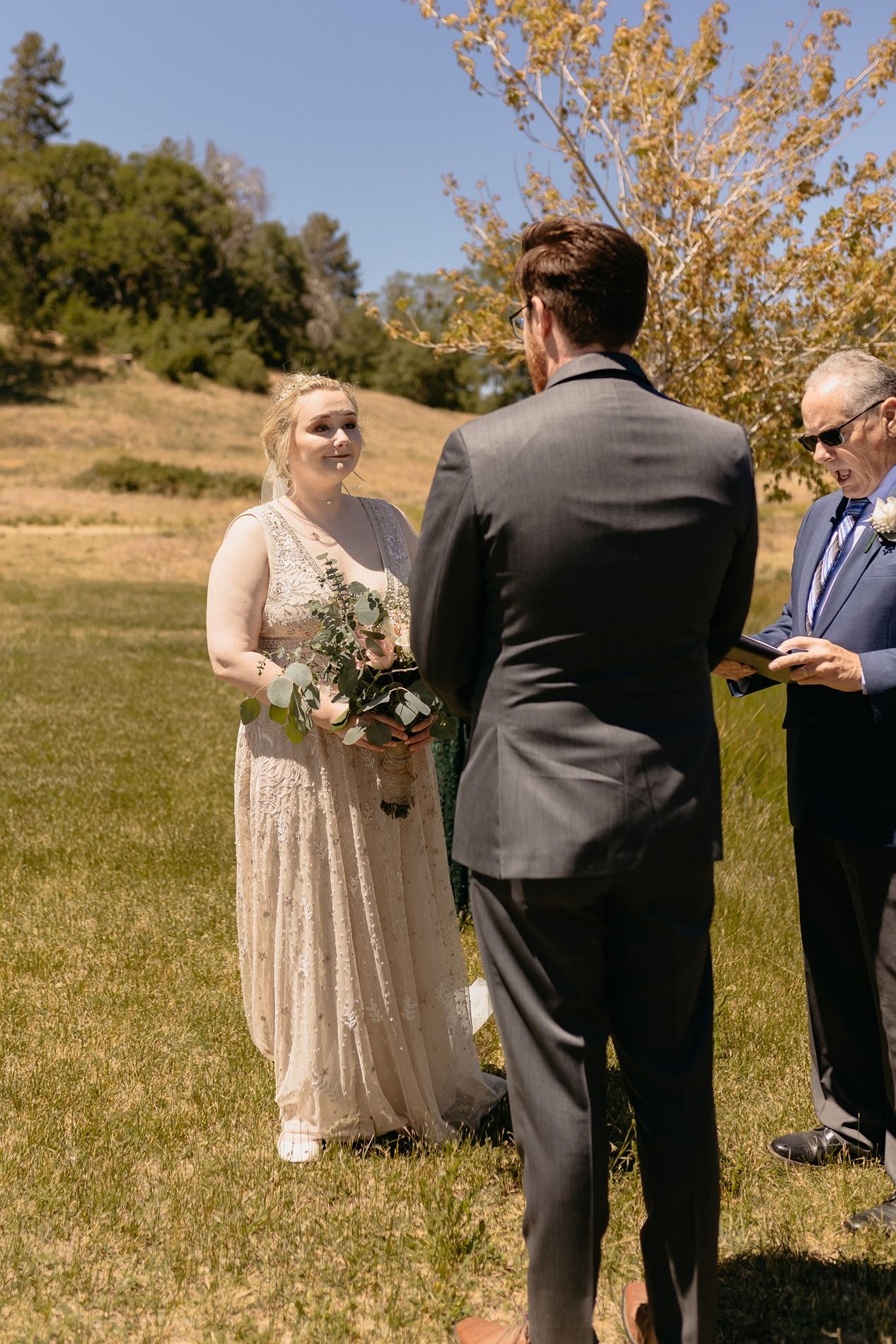 Bride in lace gown holds bouquet facing groom in gray suit during outdoor wedding ceremony on sunny day at Lake Cuyamaca, Julian.